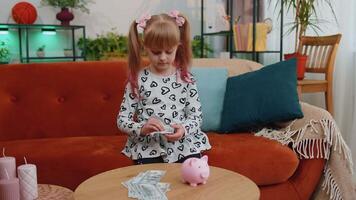 Little toddler kid girl putting money dollar banknotes for future gift purchase at piggybank at home video
