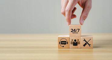 Hand placing a wooden block with 24 7 Service text on top of other blocks featuring icons of customer support, teamwork, and technical service, representing continuous business support photo