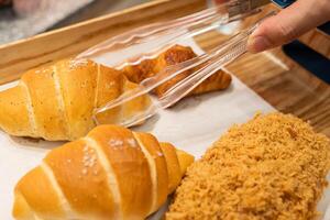 Fresh Bakery Bread Selection with Tongs in a Wooden Tray. variety of fresh bakery bread placed on a wooden tray. A hand is using plastic tongs to pick up. bakery themes in commercial or editorial use. photo