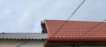 A man climbed onto the roof examining the gable at the junction of the multi-level roofs of a village house. Mobile photo