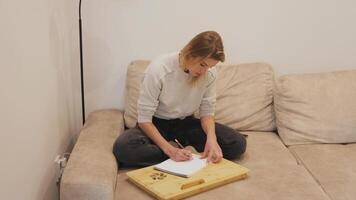 young woman sitting cross-legged on comfortable sofa, wearing casual clothes and drawing with focused concentration using lap desk and notebook video