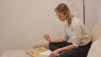 young woman sits on a couch, using a lap desk to draw and meticulously fold a piece of paper, engrossed in her activity video