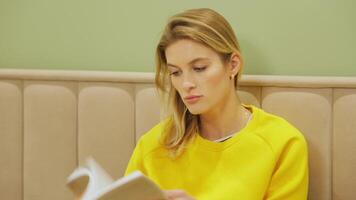 young woman reading book while sitting cross-legged on soft bed, wearing yellow sweater and gray jeans in peaceful bedroom with warm atmosphere video