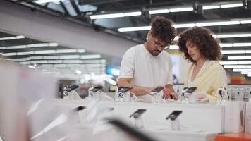 Young couple carefully selecting newest smartphone models, comparing features and technologies while browsing electronic store display in slow-motion contemplation video