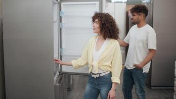 Young customers are choosing a new refrigerator in an electronics store, opening and closing its door, evaluating its features and discussing their choice video