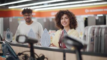 Customers are choosing a new iron in a home appliance store, a man and a woman are holding irons in their hands and smiling, looking at each other video