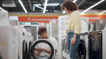 Couple shopping for washing machine, partner highlighting key features while discussing options in electronics store, capturing decision-making process in slow motion video