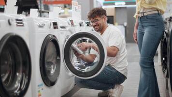 Young couple browsing home appliance store, comparing washing machine features with cheerful interaction, exploring options for domestic purchase in slow-motion setting video
