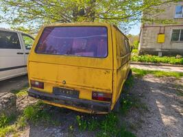 A yellow van is parked in a grassy area. The van is old and dirty, and it is parked next to a building photo