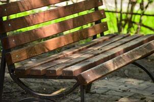 A wooden bench with a metal frame sits in a park. The bench is empty and the sun is shining on it photo