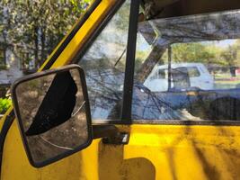 A yellow van with a broken mirror. The reflection of a white car can be seen in the rearview mirror photo
