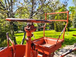 An old red machine with a rusty handle and a red tray. The machine is in a park with trees and grass photo