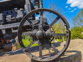 A black wheel of an old tractor is on a rock. The wheel is rusted and has a worn appearance. The scene is set in a park, with a few people in the background photo
