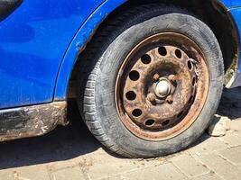 A rusted tire on a car. The tire is black and has a white rim. The tire is old and worn, and it is dirty. The car is parked on a street, and the tire is in the foreground photo