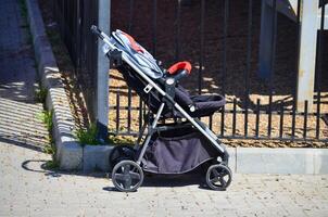 A stroller is sitting on the sidewalk next to a fence. The stroller is black and red photo