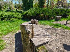 A wooden bench with a slanted top sits in a park. The bench is surrounded by trees and grass, creating a peaceful and serene atmosphere. The bench appears to be a simple and rustic design photo
