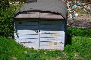 A small, old blue garage with a roof. The garage is covered in rust and has a door that is open. The garage is surrounded by grass and weeds photo