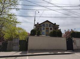 A large brown house with a white fence and a black gate. The house is surrounded by trees and has a lot of wires above photo