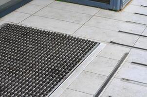 A metal grate with a black and silver design sits on a gray tile floor. The grate is positioned in front of a door, and the tile floor is tiled in a pattern. Concept of order and structure photo