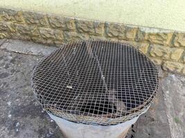 A metal trash can with a mesh lid is sitting on a sidewalk. The mesh lid is damaged and the trash can is currently empty photo