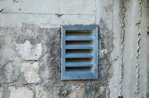 A vent is on a wall. The vent is open and has a metal grate. The vent is on a wall that is dirty and has some cracks photo