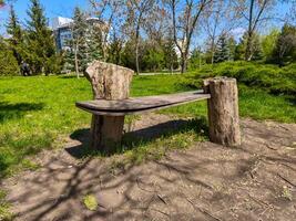 A wooden bench is sitting on a dirt field. The bench is made of wood and has a slanted back. The bench is surrounded by grass and trees photo