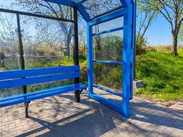 A blue bus stop with a bench and a door. The bench is empty. The sky is blue and the grass is green photo