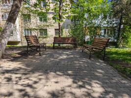 Three wooden benches are placed in a park under a tree. The park is empty and quiet, with no people in sight. The benches are arranged in a row, with one on the left, one in the middle photo