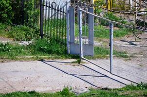 A gate is open in a yard with a fence behind it. The gate is made of metal and has a lock on it. The yard is overgrown with grass and weeds photo