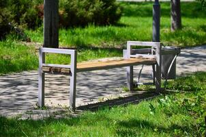 A park bench is sitting in the grass next to a trash can. The bench is empty and the grass is lush and green photo