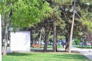 A park with a white building and a bench. The park is empty except for a few cars parked in the background photo