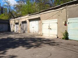 A row of old, abandoned garages sit empty in a parking lot. The buildings are grey and the sky is blue photo