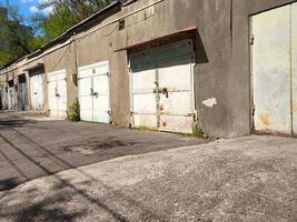 A row of old, abandoned garages sit empty on a street. The buildings are grey and the sky is blue photo