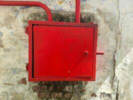 A red metal box with a handle sits on a wall. The box is made of metal and has a red color photo