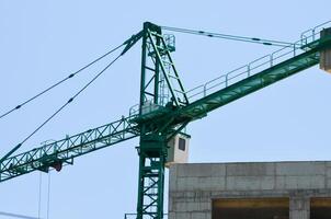 A large green crane is in the air above a building. The crane is tall and has a lot of metal parts. The sky is clear and blue, and the crane is the main focus of the image photo