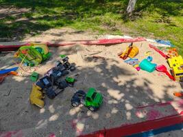 A pile of toys is scattered on the ground in a park. The toys include a yellow toy truck, a green toy truck, and a red toy truck. There are also a few other toys, such as a yellow toy car photo