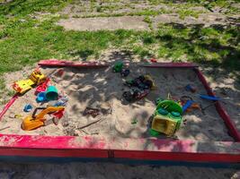 A sandbox with a variety of toys scattered around it. The toys include a green toy car, a yellow toy truck, and a blue toy truck. The sandbox is empty, but it is a fun photo