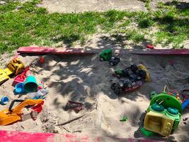 A pile of toys is scattered on the ground in a sandy area. The toys include a green truck, a red truck, a yellow truck, and a blue truck. There are also a few other toys, such as a toy car photo