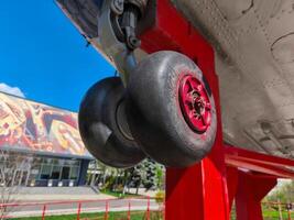 A red and white airplane with its landing gear down. The landing gear is black and red, and it is attached to a red post. The scene is set in a courtyard, with a building in the background photo