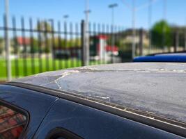A car with a dirty windshield sits in a parking lot. The car is parked next to a fence and there is a soccer field in the background photo