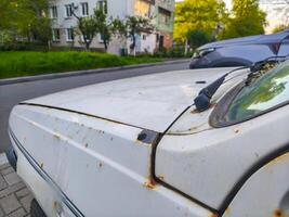 A white car with a rusted hood and a dirty windshield. The car is parked on a street next to a building photo