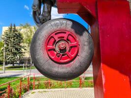 A red wheel with black rubber on it is hanging from a red pole. The wheel is in a park with a fence and a building in the background photo