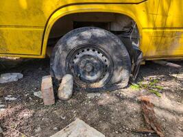 A yellow bus with a black tire on the ground. The tire is missing and there is a brick on the ground next to it photo