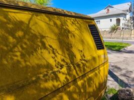 A yellow van is parked on the side of the road. The van is covered in dirt and has a lot of leaves on it. The leaves are casting a shadow on the van, making it look even more dirty. The scene is quiet photo