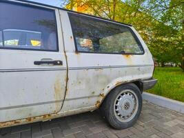 A white car with rusted metal parts and a faded blue stripe. The car is parked on a brick road photo