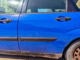 A blue car with dirty tires and a black stripe on the side. The car is parked in front of a building photo