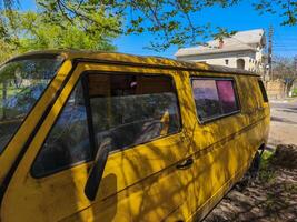 A yellow van is parked in a grassy area. The van is old and has a faded yellow color. The windows are open, and the van is parked in the shade photo