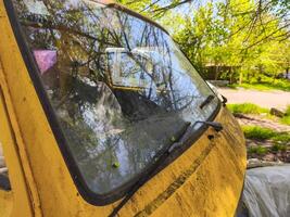 A yellow car with a dirty windshield and wipers. The windshield is covered in dirt and the wipers are covered in mud. The car is parked in a grassy area photo