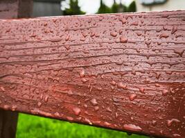The bench is wet with raindrops on it. The bench is made of wood and has a reddish color photo