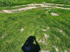 A man takes a picture of a grassy field. The grass is green, the field is empty and only a shadow is visible from the man. photo